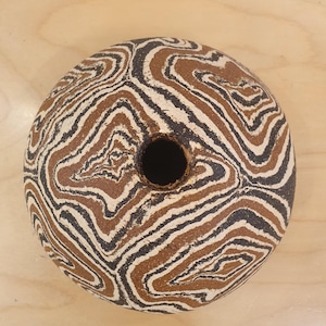 May include: A spherical ceramic vase featuring a marbled pattern of swirling black, white, and earthy brown clay layers. The object has a circular opening at the top center, showcasing a textured, matte finish across its rounded, organic form.