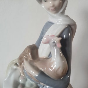 Pode incluir: Uma figura de porcelana de uma mulher usando um len&ccedil;o branco na cabe&ccedil;a e um vestido azul, segurando um frango branco nos bra&ccedil;os. Ela est&aacute; carregando uma cesta de vime com um forro de tecido branco.
