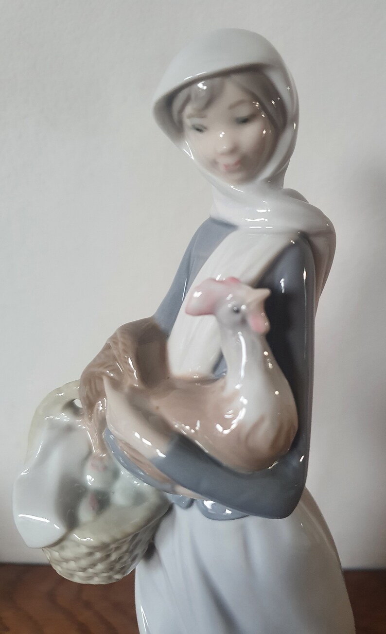Pode incluir: Uma figura de porcelana de uma mulher usando um len&ccedil;o branco na cabe&ccedil;a e um vestido azul, segurando um frango branco nos bra&ccedil;os. Ela est&aacute; carregando uma cesta de vime com um forro de tecido branco.