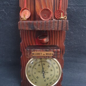 May include: A wooden wall thermometer with a round face and a gold trim. The thermometer has a scale in both Fahrenheit and Celsius. The thermometer is mounted on a wooden plaque with a carved design. The plaque has a small shelf with a copper pot and a sign that reads "LLORET de MAR".