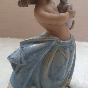 May include: A ceramic figurine of a girl with long brown hair, wearing a blue and white dress, holding a white pitcher. The figurine is standing on a wooden base.