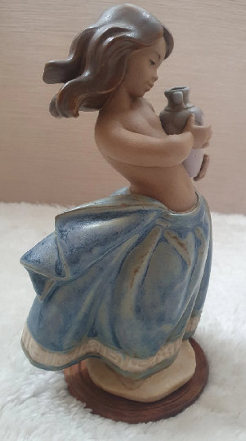 May include: A ceramic figurine of a girl with long brown hair, wearing a blue and white dress, holding a white pitcher. The figurine is standing on a wooden base.