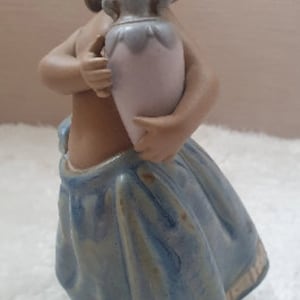 May include: A porcelain figurine of a girl wearing a blue dress and holding a light purple vase.