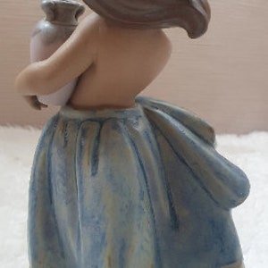 May include: A ceramic figurine of a girl with brown hair wearing a blue and white dress and holding a ceramic jug.