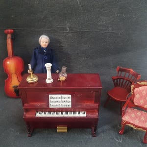 May include: A miniature dollhouse scene with a red piano, two chairs, a doll, a cello, and a lamp. The piano has a music sheet with notes on it.