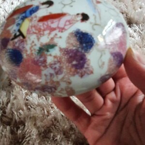 May include: A white porcelain trinket box with a floral design and a scene of people in traditional clothing. The box is decorated with pink, red, blue, and purple flowers.