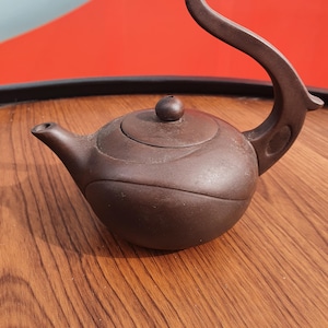 May include: A dark brown teapot with a matte finish, featuring a rounded body, a short spout, and a high, curved handle that extends upward. The lid is circular with a small knob at the center, and the body has subtle, flowing curved lines.