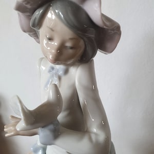 May include: A porcelain figurine of a girl wearing a pink hat and a white dress, holding a white bird in her hands.