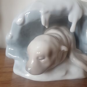May include: A ceramic figurine depicting a polar bear cub in an ice cave. The sculpture shows a white bear with a grey nose and closed eyes, set against a light blue and white ice backdrop. The figurine has a glossy finish.