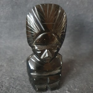 May include: Black obsidian sculpture of a stylized human figure with a tall, pointed headdress. The sculpture is carved with intricate details and has a smooth, polished finish.