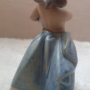 May include: A ceramic figurine of a woman with brown hair wearing a blue and white dress. The figurine is standing on a wooden base.
