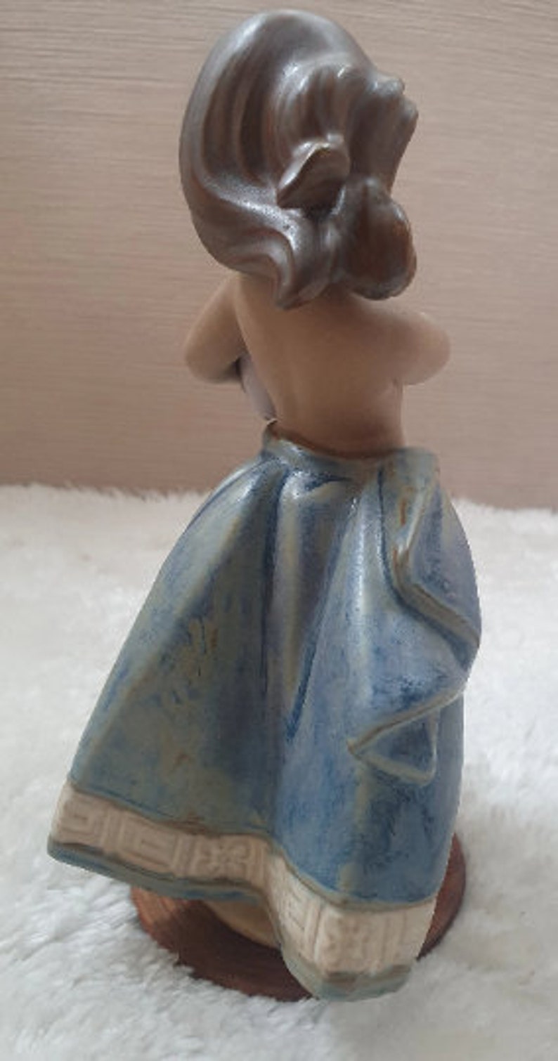 May include: A ceramic figurine of a woman with brown hair wearing a blue and white dress. The figurine is standing on a wooden base.