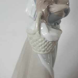 Pode incluir: Uma figura de porcelana de uma mulher vestindo um vestido branco com capuz e segurando uma cesta com um frango dentro. A figura &eacute; pintada em tons de branco, azul e marrom.