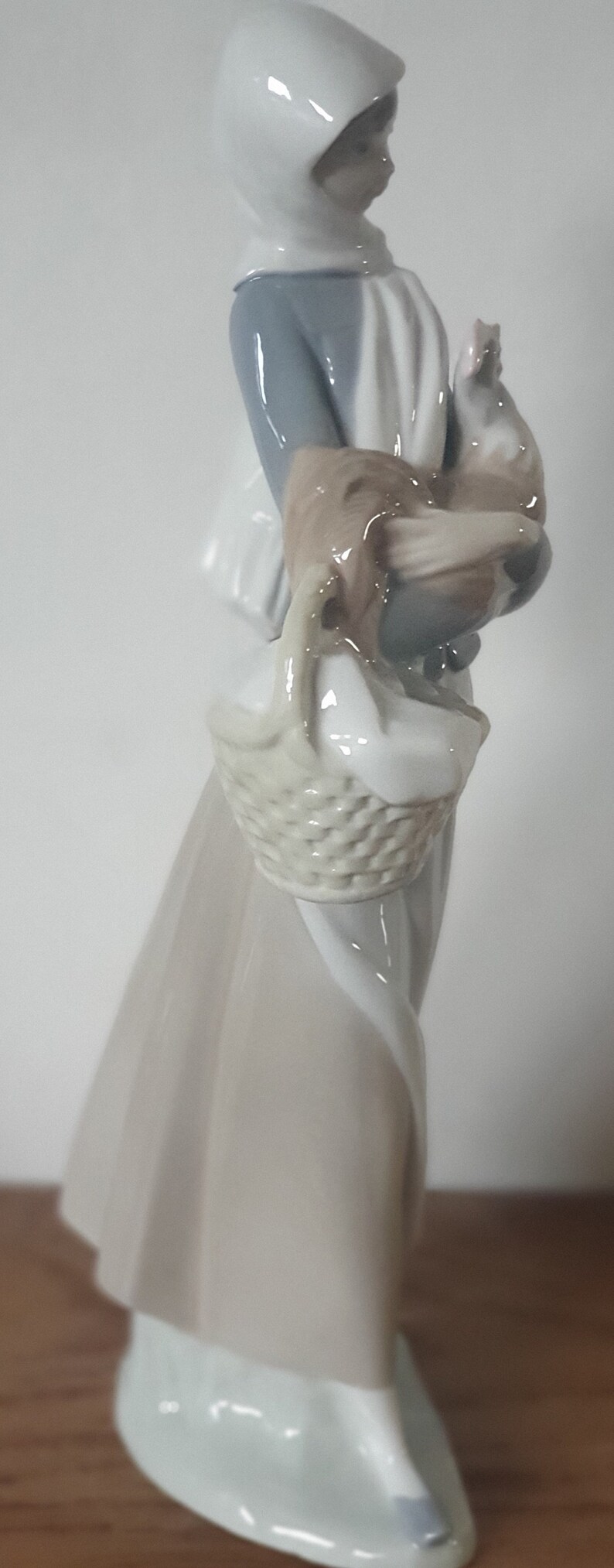 Pode incluir: Uma figura de porcelana de uma mulher vestindo um vestido branco com capuz e segurando uma cesta com um frango dentro. A figura &eacute; pintada em tons de branco, azul e marrom.