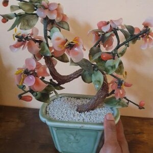 May include: A decorative bonsai tree sculpture featuring carved pink stone flowers and translucent green stone leaves attached to dark brown branches. The tree is planted in a square, light green ceramic pot filled with small white gravel.