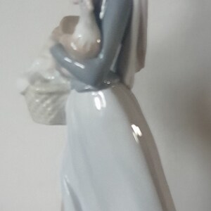 Pode incluir: Uma figura de porcelana de uma mulher com um vestido azul e um len&ccedil;o branco na cabe&ccedil;a segurando um frango branco nos bra&ccedil;os. Ela est&aacute; carregando uma cesta na outra m&atilde;o.