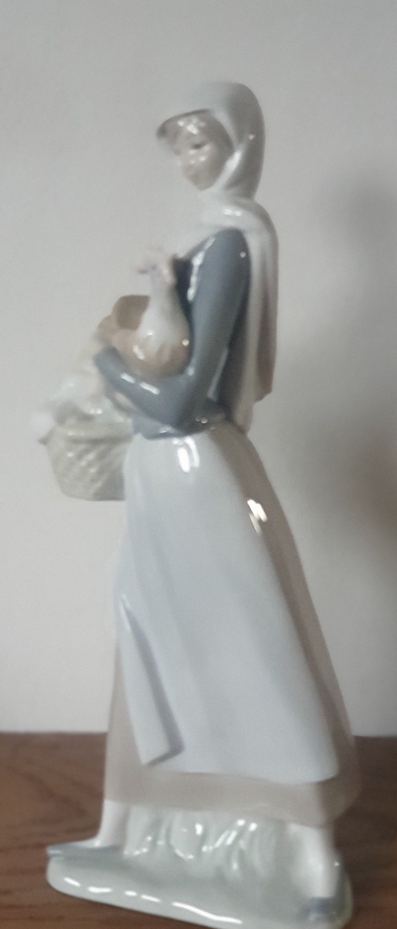 Pode incluir: Uma figura de porcelana de uma mulher com um vestido azul e um len&ccedil;o branco na cabe&ccedil;a segurando um frango branco nos bra&ccedil;os. Ela est&aacute; carregando uma cesta na outra m&atilde;o.