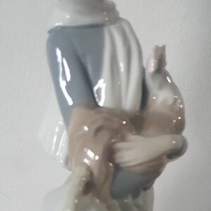 Pode incluir: Uma figura de porcelana de uma menina vestindo um casaco branco com capuz e segurando um p&aacute;ssaro marrom e branco. A menina est&aacute; vestindo um vestido azul e um cachecol branco. Ela est&aacute; segurando uma cesta na outra m&atilde;o.