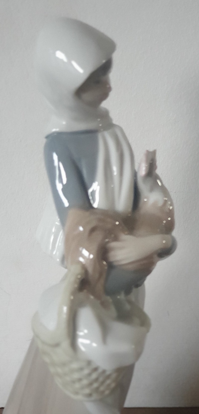 Pode incluir: Uma figura de porcelana de uma menina vestindo um casaco branco com capuz e segurando um p&aacute;ssaro marrom e branco. A menina est&aacute; vestindo um vestido azul e um cachecol branco. Ela est&aacute; segurando uma cesta na outra m&atilde;o.
