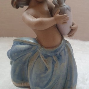 May include: A ceramic figurine of a young girl with brown hair, wearing a blue dress and holding a light purple pitcher.