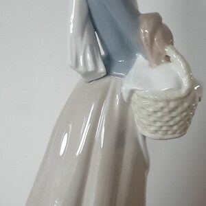 Pode incluir: Uma figura de porcelana de uma mulher usando um len&ccedil;o branco na cabe&ccedil;a e um vestido azul, segurando uma cesta e um pequeno p&aacute;ssaro marrom.