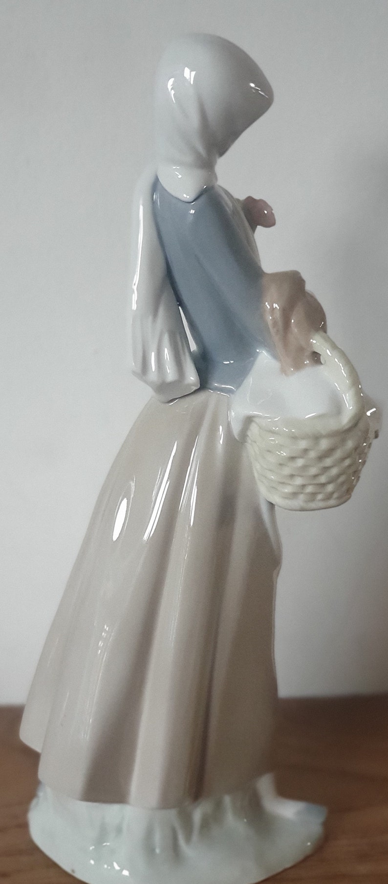 Pode incluir: Uma figura de porcelana de uma mulher usando um len&ccedil;o branco na cabe&ccedil;a e um vestido azul, segurando uma cesta e um pequeno p&aacute;ssaro marrom.
