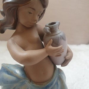 May include: A ceramic figurine of a young girl with brown hair holding a small, light purple vase. The girl is wearing a blue and white dress.
