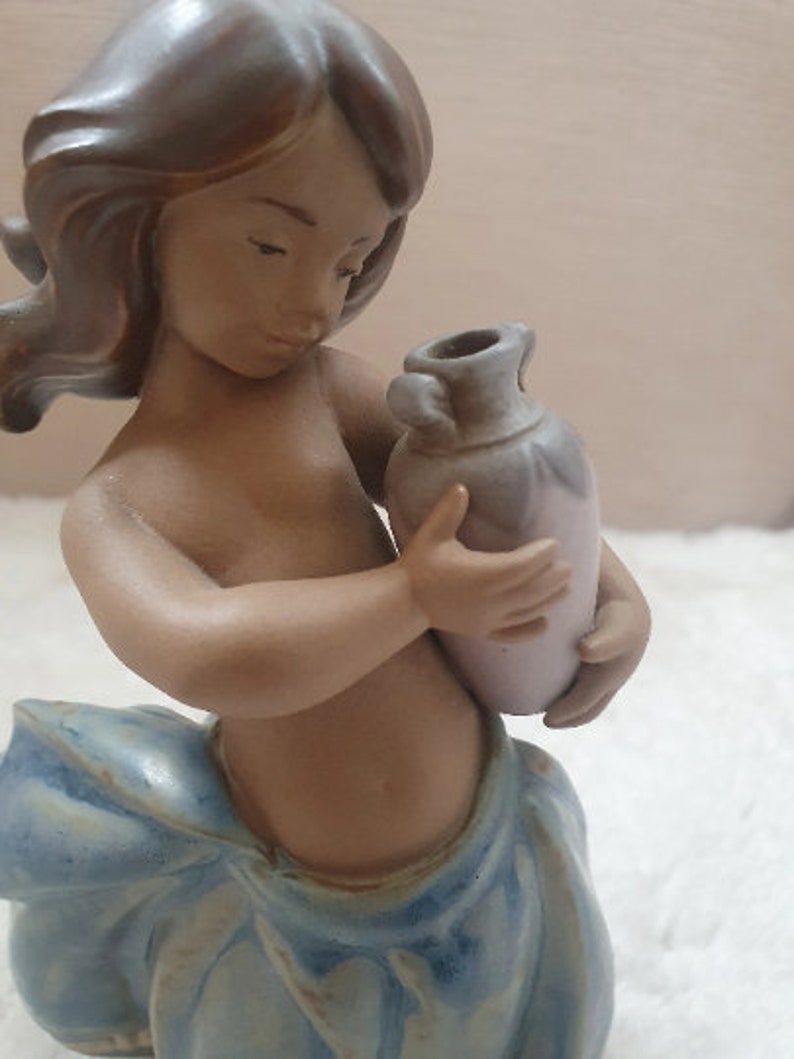 May include: A ceramic figurine of a young girl with brown hair holding a small, light purple vase. The girl is wearing a blue and white dress.