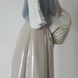 Pode incluir: Uma figura de porcelana de uma mulher usando um chap&eacute;u branco e um vestido azul, segurando uma cesta de vime. A figura est&aacute; em p&eacute; sobre uma superf&iacute;cie de madeira.