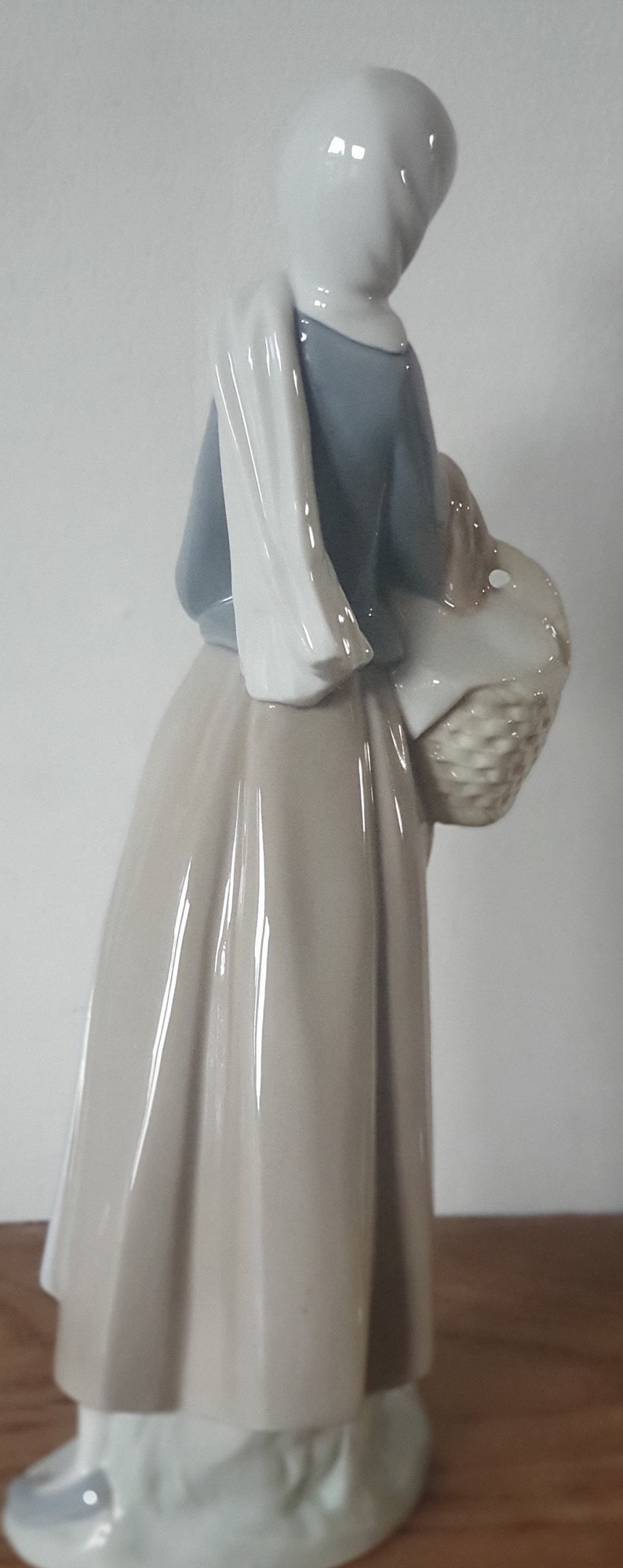 Pode incluir: Uma figura de porcelana de uma mulher usando um chap&eacute;u branco e um vestido azul, segurando uma cesta de vime. A figura est&aacute; em p&eacute; sobre uma superf&iacute;cie de madeira.