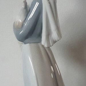 Pode incluir: Uma figura de porcelana de uma mulher vestindo um manto branco com capuz e um vestido azul, segurando um pequeno p&aacute;ssaro branco em seus bra&ccedil;os.