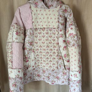Puede incluir: Chaqueta acolchada con diseño patchwork. La chaqueta tiene capucha y bolsillo delantero. La tela presenta estampados rosas y blancos, incluyendo estampados florales y cuadros vichy. La chaqueta está colgada en una percha.