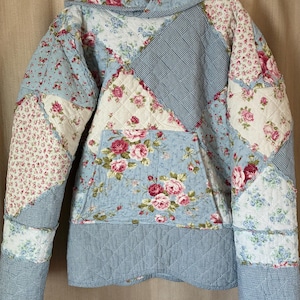 May include: A quilted jacket with a patchwork design. The jacket features a hood and a front pocket. The fabric is a mix of floral patterns in pink and red on a light blue background, with sections of blue and white gingham.