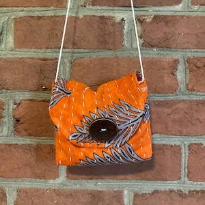 May include: A small orange and gray patterned fabric bag with a wooden button closure. The bag is hanging from a white string.