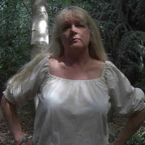 May include: A cream-coloured peasant blouse with a square neckline and elbow-length sleeves. The blouse is made of a lightweight fabric and has a gathered detail at the neckline and sleeves. The person is wearing a turquoise bracelet.