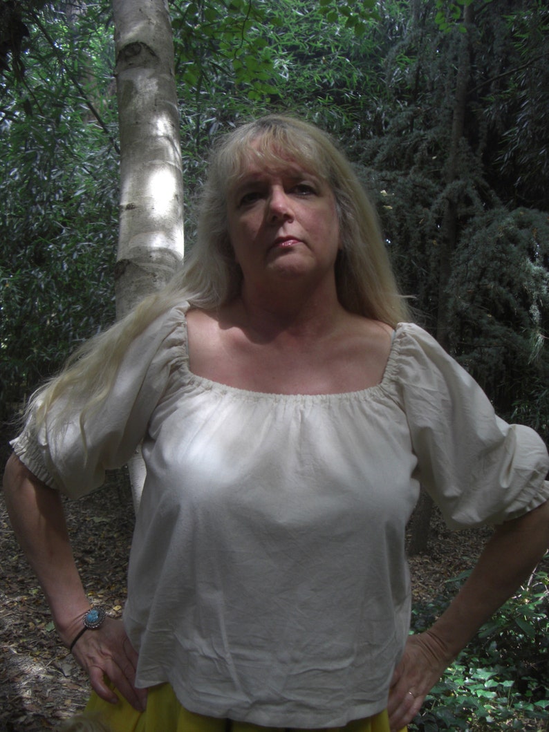 May include: A cream-coloured peasant blouse with a square neckline and elbow-length sleeves. The blouse is made of a lightweight fabric and has a gathered detail at the neckline and sleeves. The person is wearing a turquoise bracelet.
