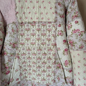 Könnte beinhalten: Gesteppte Jacke mit Patchwork-Design. Die Jacke hat verschiedene Blumenmuster in Rosa und Weiß, mit hellrosa Vichy-Stoff auf der Kapuze. Die Jacke hat eine Tasche und eine gewellte Bordüre.
