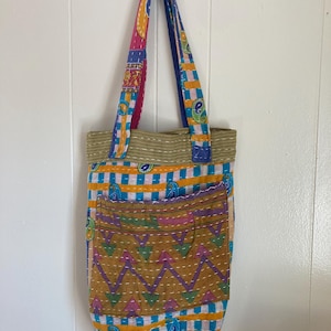 May include: A colorful tote bag with a patchwork design. The bag features a variety of patterns, including stripes, paisley, and chevron. The bag has a front pocket and two handles. The colors include blue, yellow, pink, and green.