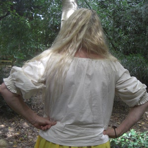 May include: A cream-coloured peasant blouse with three-quarter sleeves. The blouse features a gathered neckline and cuffs, and a slightly puffed sleeve design. The fabric appears to be a lightweight cotton or linen. The background is a natural outdoor setting with green foliage.