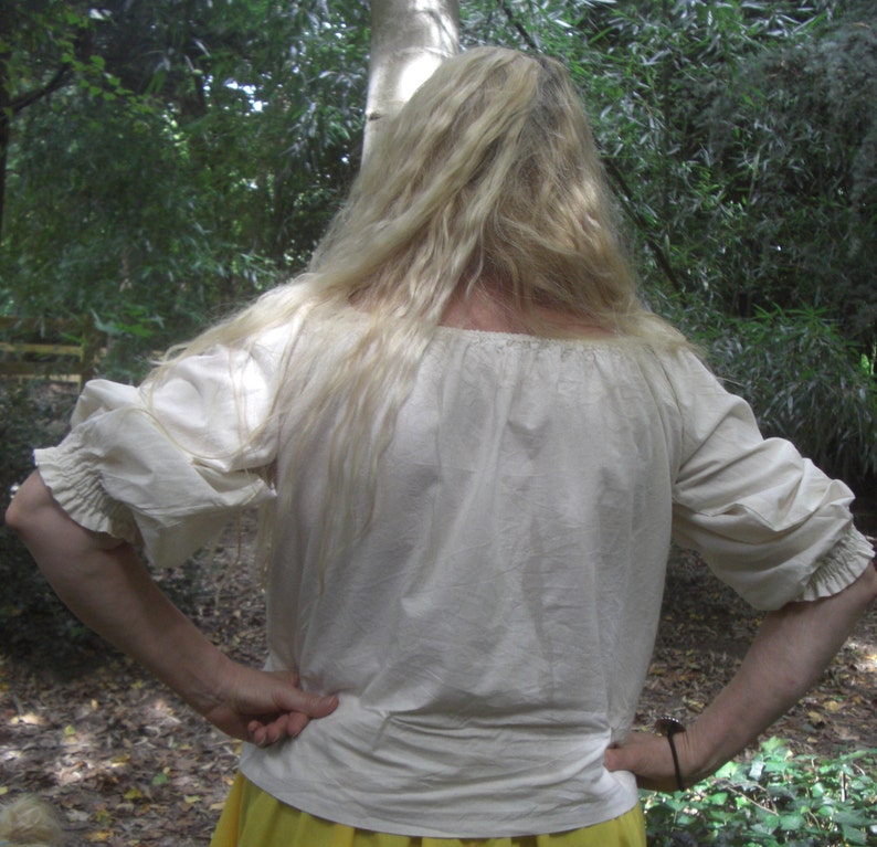 May include: A cream-coloured peasant blouse with three-quarter sleeves. The blouse features a gathered neckline and cuffs, and a slightly puffed sleeve design. The fabric appears to be a lightweight cotton or linen. The background is a natural outdoor setting with green foliage.