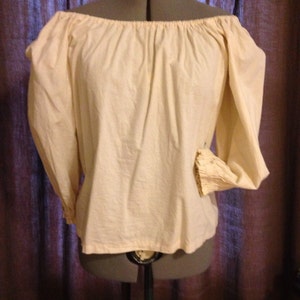 May include: Cream-coloured off-the-shoulder blouse with long sleeves. The blouse is made of a soft, slightly textured fabric. The sleeves have a gathered detail at the cuffs. The blouse is displayed on a mannequin.
