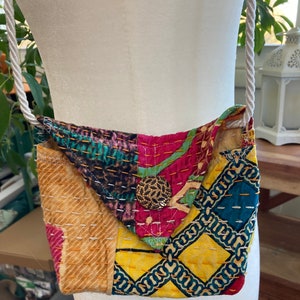 May include: A small, colorful patchwork bag with a white rope strap. The bag is made of different fabrics with a variety of patterns, including floral, geometric, and paisley. The bag has a single button closure.
