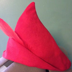 May include: A red felt elf hat with a pointed tip and a feather attached to the front.