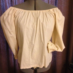 May include: Cream-coloured off-the-shoulder blouse with long, puffy sleeves. The sleeves gather at the wrists with a ruffled cuff. The blouse is displayed on a dress form against a dark curtain backdrop.