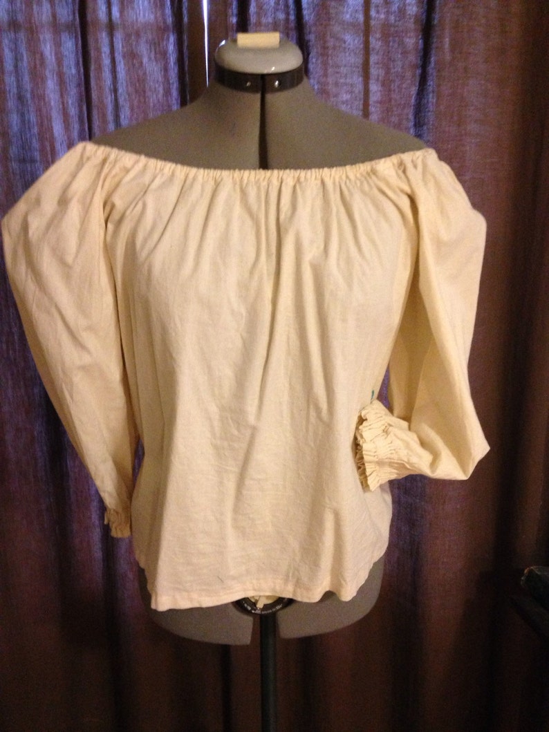 May include: Cream-coloured off-the-shoulder blouse with long, puffy sleeves. The sleeves gather at the wrists with a ruffled cuff. The blouse is displayed on a dress form against a dark curtain backdrop.