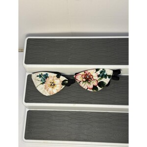 May include: Two floral eye masks with black elastic bands. The masks feature a cream-colored background with pink, peach, and green floral designs. The eye masks are designed to block out light.