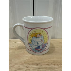 May include: White ceramic mug with a cartoon doll illustration in a fur coat and purse. The mug has a pink border and floral accents. The handle is curved. The mug is on a wooden surface.