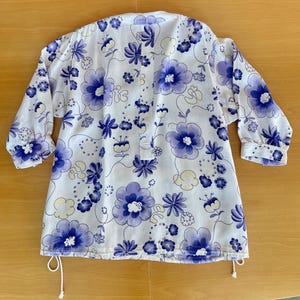 May include: White tunic with a floral pattern in shades of purple and blue. The tunic has long sleeves with buttoned cuffs and a drawstring at the hem. The floral design includes large and small flowers and leaves.