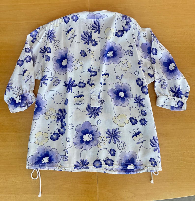 May include: White tunic with a floral pattern in shades of purple and blue. The tunic has long sleeves with buttoned cuffs and a drawstring at the hem. The floral design includes large and small flowers and leaves.