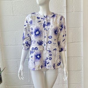May include: White blouse with a blue floral pattern. The blouse has a round collar, three-quarter sleeves, and a button-down front. The floral design features large and small blue flowers with yellow accents.